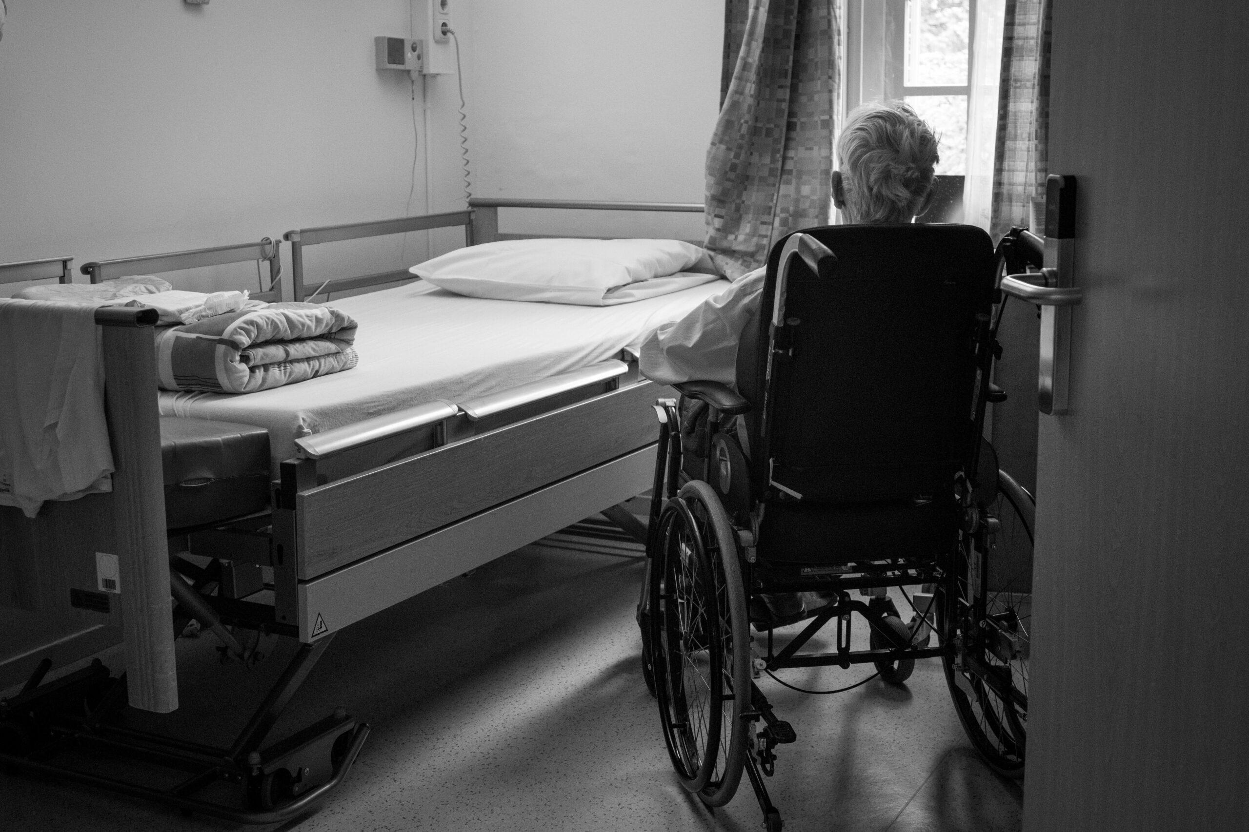 An elderly person in a wheelchair facing an empty hospital bed in a quiet, dimly lit room, with soft daylight coming through the window and folded linens placed neatly on the mattress.