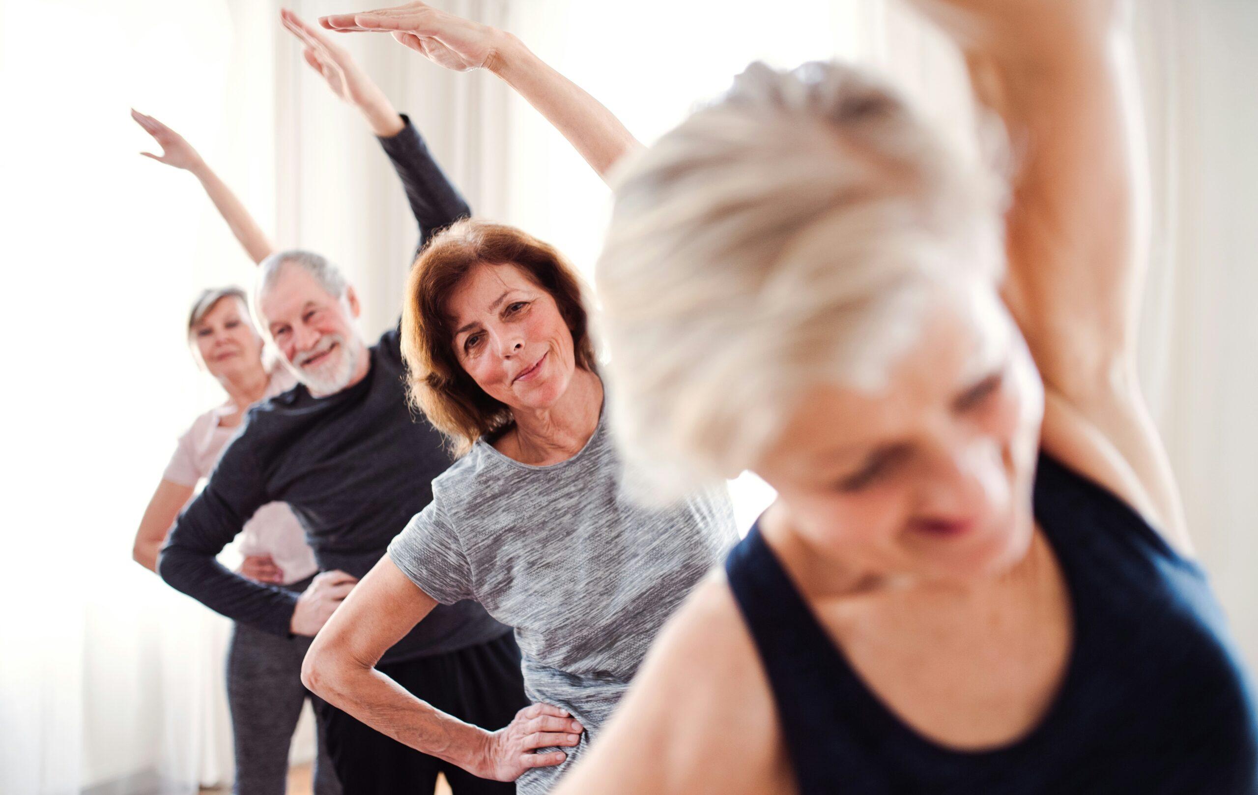 A group of older adults in workout clothing standing in a row indoors, smiling as they perform a side-stretch exercise with one arm raised overhead.