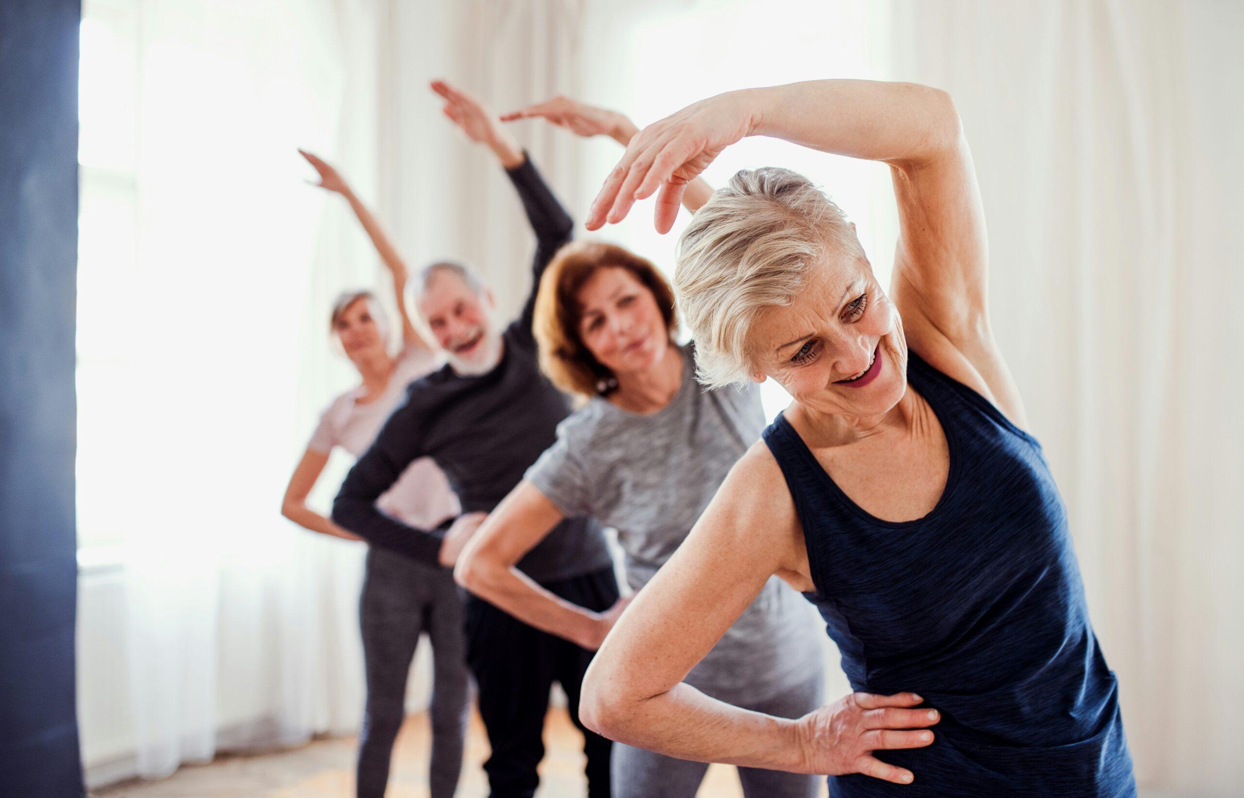 A group of older adults standing in a bright studio, smiling as they perform coordinated side-stretch exercises with one arm raised overhead, appearing relaxed, energetic, and engaged in a fitness class together.