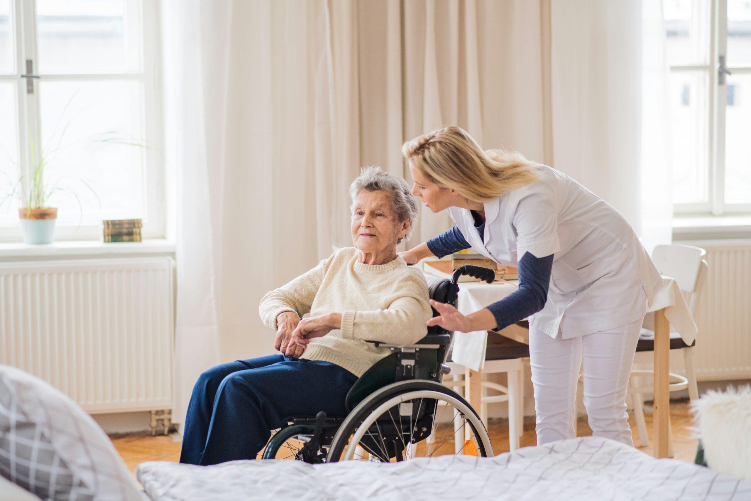 An elderly woman sitting in a wheelchair beside a bright window while a caregiver in white medical attire leans in supportively, resting a hand on the chair and speaking gently to her in a calm, home-like room.