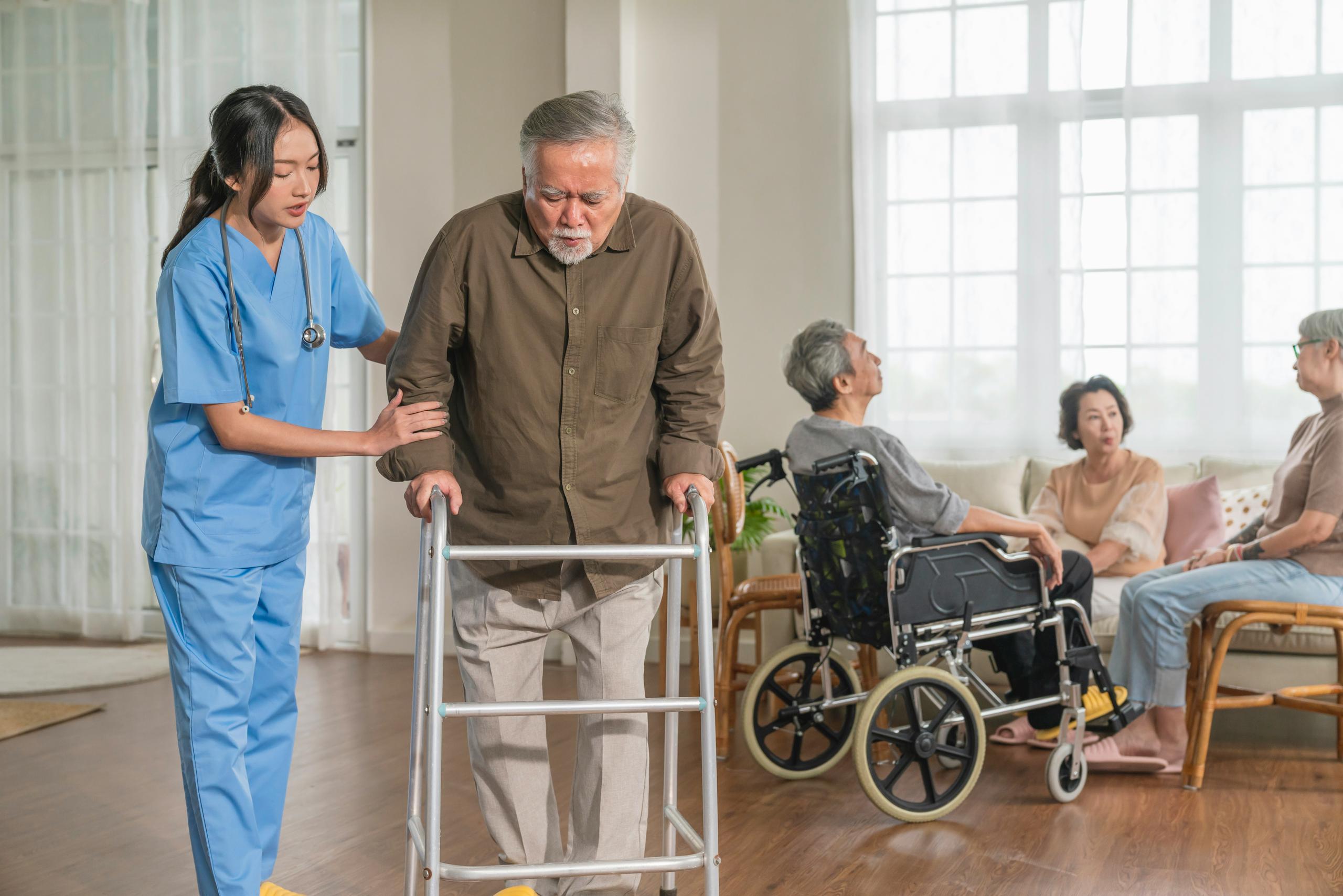 An older man using a walker for support while a nurse in blue scrubs stands beside him offering assistance, with other elderly individuals seated and talking in the background of a bright care-facility room.