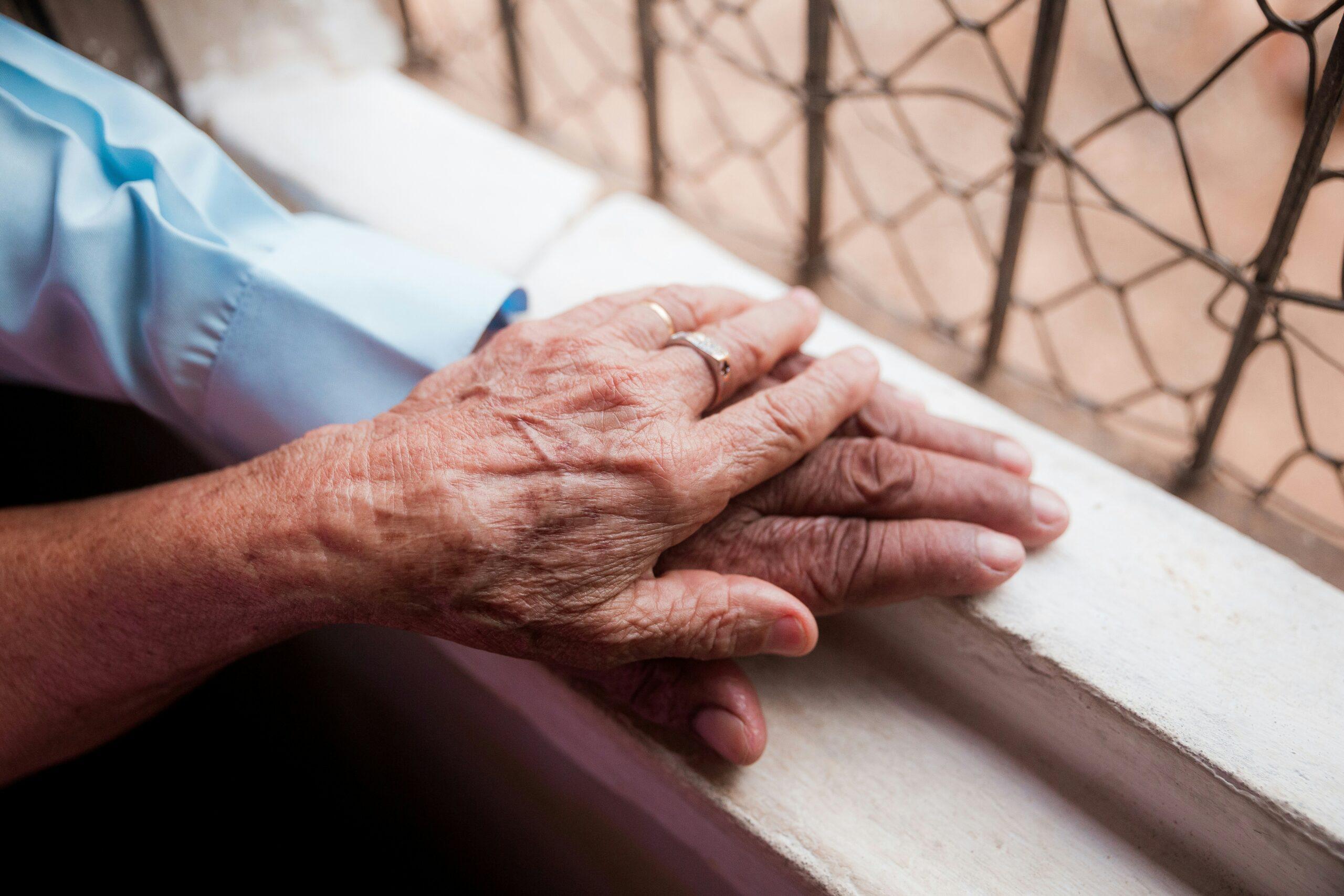 Two elderly, wrinkled hands gently resting on top of each other on a windowsill, softly lit by daylight, with a wire-patterned window in the background creating an intimate, quiet moment of support and closeness.
