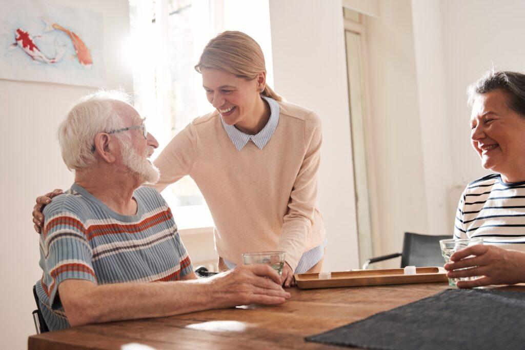 A smiling caregiver stands beside an older man and woman at a wooden table, gently placing a hand on the man’s shoulder as the group shares a warm moment together, holding drinks in a bright, cozy room.
