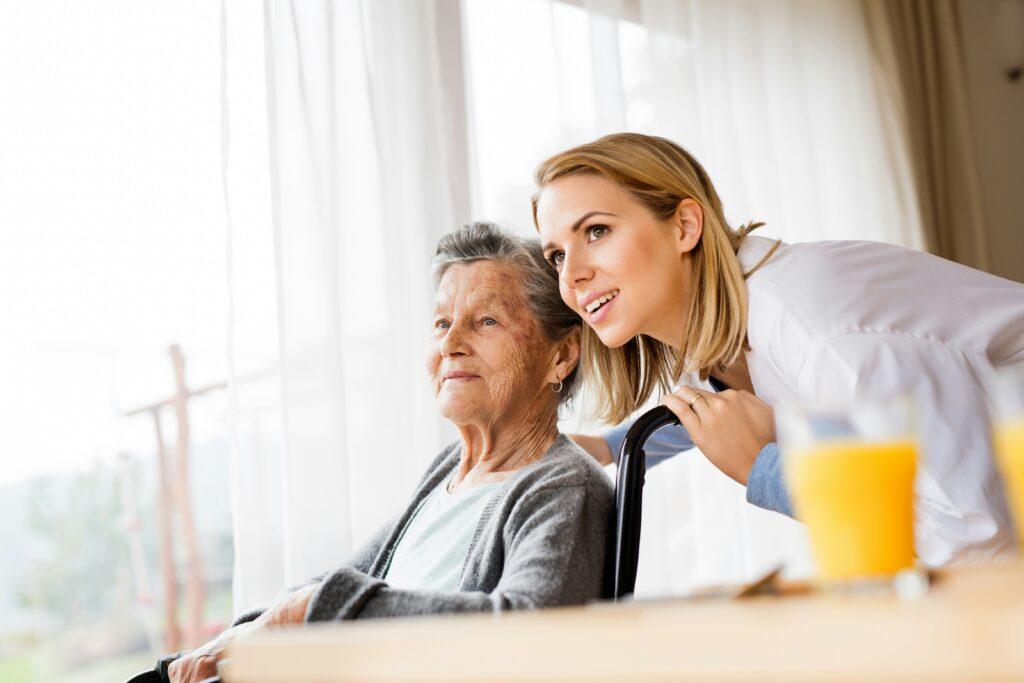 Smiling elderly woman seated indoors beside a large window with sheer curtains, while a younger caregiver in a white coat leans in close behind her wheelchair, both looking toward the light, with a glass of orange juice blurred in the foreground, conveying warmth, support, and attentive care in a calm, home-like setting.