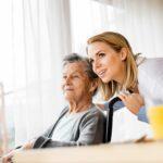 Smiling elderly woman seated indoors beside a large window with sheer curtains, while a younger caregiver in a white coat leans in close behind her wheelchair, both looking toward the light, with a glass of orange juice blurred in the foreground, conveying warmth, support, and attentive care in a calm, home-like setting.