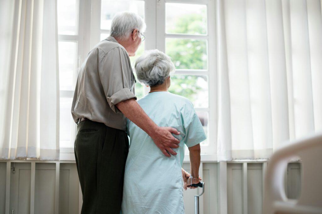 Elderly couple standing together in front of a large window, smiling and enjoying each other's company.