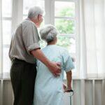Elderly couple standing together in front of a large window, smiling and enjoying each other's company.