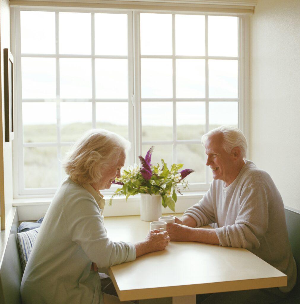 An elderly couple with gray hair sit across from each other at a small kitchen table by a large window, smiling and talking while holding cups of coffee, with soft daylight filling the room and a vase of flowers placed between them on the table.