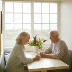 An elderly couple with gray hair sit across from each other at a small kitchen table by a large window, smiling and talking while holding cups of coffee, with soft daylight filling the room and a vase of flowers placed between them on the table.