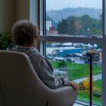 An elderly person with short curly gray hair sits in a cushioned chair by a large window, looking out at a rainy suburban landscape, with raindrops on the glass and a purple cane resting upright beside the chair.