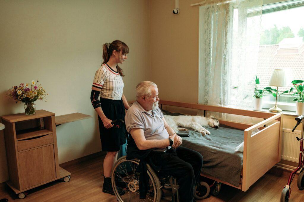A woman in a wheelchair sits beside an elderly man lying in bed, sharing a moment of connection and care.