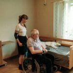 A woman in a wheelchair sits beside an elderly man lying in bed, sharing a moment of connection and care.